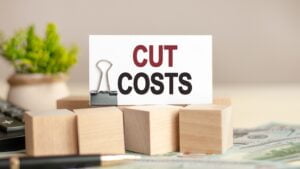 Piece of paper with the text: CUT COSTS. Wooden cubes, banknotes, pen, black calculator and green plant in the background. Business and finance concept.