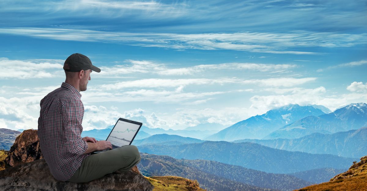 Portrait of a guy sitting at the top of a mountain working with laptop.