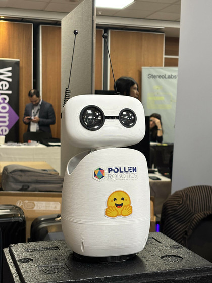 A compact humanoid robot, Reachy Mini, standing on a tabletop. It has a sleek white and black design with a head featuring animated antennae, articulated arms, and visible sensors, representing a lightweight, programmable platform for AI and robotics research.